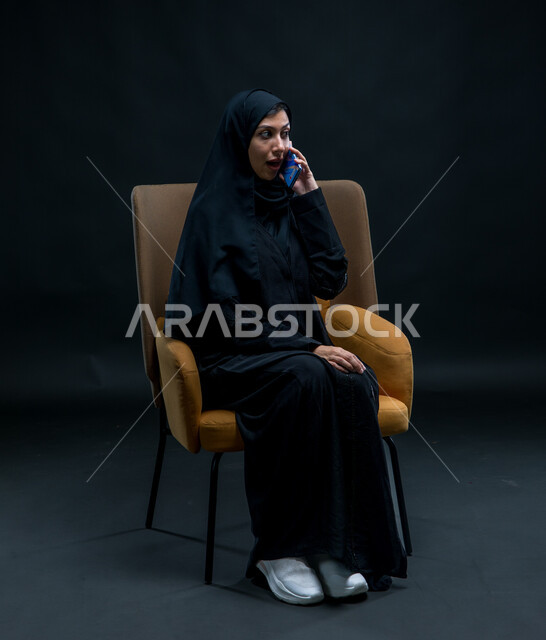 قضاء وقت الفراغ بالتواصل مع الصديقات، استخدام الأجهزة والوسائل التقنية الحديثة، إرسال الرسائل النصية والصوتية عبر الجوال، بورتريه لامرأة عربية خليجية سعودية محجبة ترتدي عباءة سوداء تجلس على كرسي مريح تقوم بإجراء مكالمة هاتفية عبر الهاتف المحمول بإيماءات الاندهاش والذهول، خلفية سوداء