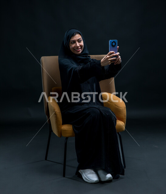 مشاركة اللحظات السعيدة على مواقع التواصل الاجتماعي، استخدام الأجهزة والوسائل التقنية الحديثة، بورتريه لامرأة عربية خليجية سعودية محجبة ترتدي عباءة سوداء تجلس على كرسي مريح تقوم بالتقاط صورة سيلفي تذكارية عبر الهاتف المحمول بإيماءات السعادة والسرور، خلفية سوداء