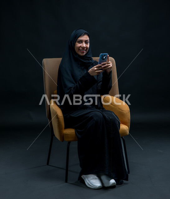 تصفح مواقع التواصل الاجتماعي، إرسال الرسائل النصية والصوتية عبر الجوال، بورتريه لامرأة عربية خليجية سعودية محجبة ترتدي عباءة سوداء تجلس على كرسي مريح تقوم باستخدام الهاتف المحمول بإيماءات السرور والاستمتاع، قضاء وقت الفراغ بالتواصل مع الصديقات، خلفية سوداء