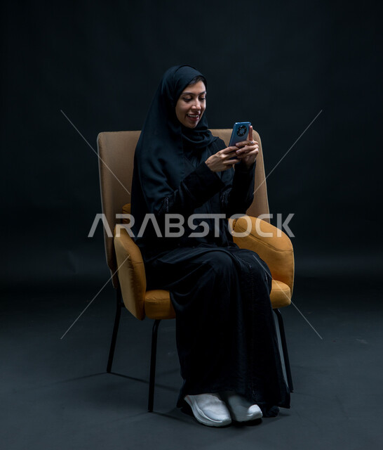 Sending text and voice messages via mobile phone, chatting and talking with family and friends, a portrait of a veiled Saudi Arabian Gulf woman wearing a black abaya sitting on a comfortable chair recording an audio clip using a mobile phone, spending free time communicating with female friends, black background