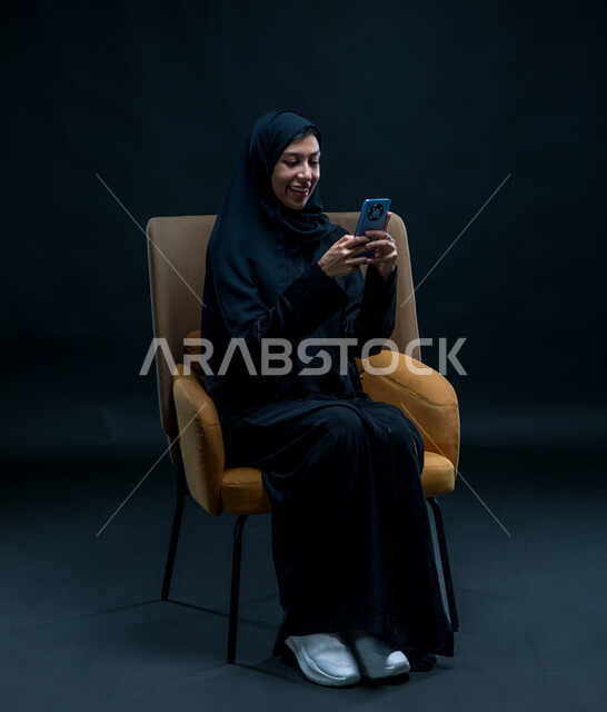 إرسال الرسائل النصية والصوتية عبر الجوال، الدردشة والتحدث مع الأهل والأصدقاء، بورتريه لامرأة عربية خليجية سعودية محجبة ترتدي عباءة سوداء تجلس على كرسي مريح تقوم بتسجيل مقطع صوتي باستخدام الهاتف المحمول، قضاء وقت الفراغ بالتواصل مع الصديقات، خلفية سوداء