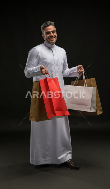 الاستفادة من عروض وتخفيضات الجمعة البيضاء، متعة التسوق واقتناء المستلزمات والهدايا، بورتريه لرجل عربي خليجي سعودي مبتسم يرتدي الثوب التقليدي يحمل بيده أكياس ورقية ملونة ينظر إلى الكاميرا بإيماءات السرور والسعادة، صورة الجسم بالطول الكامل، خلفية سوداء