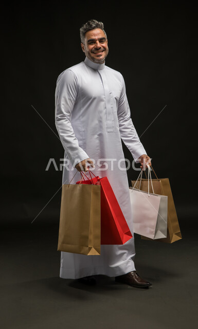 الاستفادة من عروض وتخفيضات الجمعة البيضاء، متعة التسوق واقتناء المستلزمات والهدايا، بورتريه لرجل عربي خليجي سعودي مبتسم يرتدي الثوب التقليدي يحمل بيده أكياس ورقية ملونة ينظر إلى الكاميرا بإيماءات السرور والسعادة، صورة الجسم بالطول الكامل، خلفية سوداء