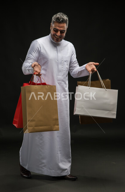 الاستفادة من عروض وتخفيضات الجمعة البيضاء، متعة التسوق واقتناء المستلزمات والهدايا، بورتريه لرجل عربي خليجي سعودي مبتسم يرتدي الثوب التقليدي يحمل بيده أكياس ورقية ملونة بإيماءات السرور والسعادة، صورة الجسم بالطول الكامل، خلفية سوداء