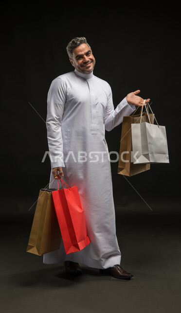 النظر إلى الكاميرا بإيماءات السرور والسعادة، بورتريه لرجل عربي خليجي سعودي مبتسم يرتدي الثوب التقليدي يحمل بيده أكياس ورقية ملونة، تعابير الحماس للتسوق واقتناء المستلزمات والهدايا، الاستفادة من عروض وتخفيضات الجمعة البيضاء، صورة الجسم بالطول الكامل، خلفية سوداء