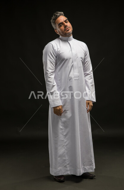الشعور بالملل والتعب من ضغوطات العمل والحياة، بورتريه لرجل عربي خليجي سعودي يرتدي الثوب التقليدي ينظر الى الكاميرا بإيماءات التعب والارهاق، مفهوم عدم القدرة على تكملة شيء ما، صورة الجسم بالطول الكامل، خلفية سوداء