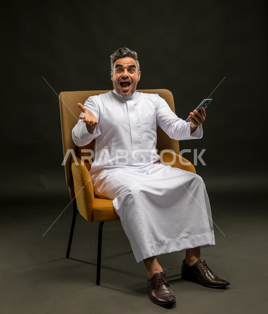 الاشارة إلى الهاتف المحمول بتعابير الانفعال والاندهاش، تصفح وسائل التواصل الاجتماعي، قضاء اوقات ممتعة باستخدام التقنيات الحديثة، بورتريه لشاب عربي خليجي سعودي يرتدي الثوب التقليدي يجلس على كرسي يحمل جوال بتعابير الذهول والسرور، خلفيه سوداء