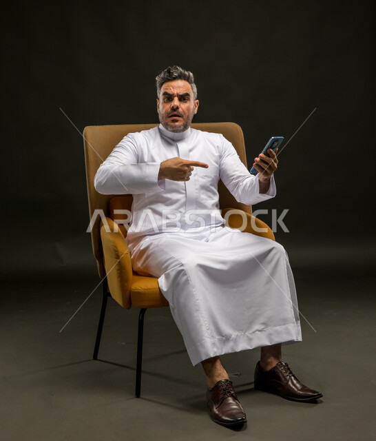الاشارة إلى الهاتف المحمول بتعابير الغضب والانزعاج، تصفح وسائل التواصل الاجتماعي، استخدام التقنيات الحديثة، بورتريه لشاب عربي خليجي سعودي يرتدي الثوب التقليدي يجلس على كرسي يحمل جوال بتعابير عدم الرضا، خلفيه سوداء