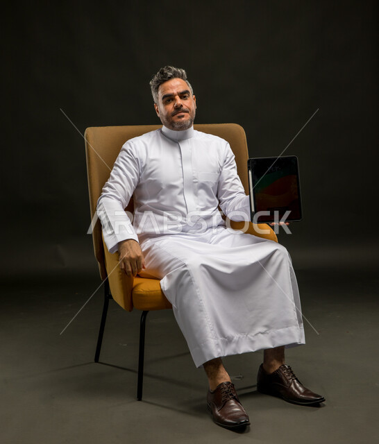 النظر إلى الكاميرا بإيماءات الانزعاج، استخدام الأجهزة والتقنيات الحديثة، التسويق الرقمي الالكتروني، عرض شاشة إعلانية سوداء فارغة، بورتريه لشاب عربي خليجي سعودي يرتدي الزي التقليدي يمسك  بيده جهاز التابلت، خلفية سوداء