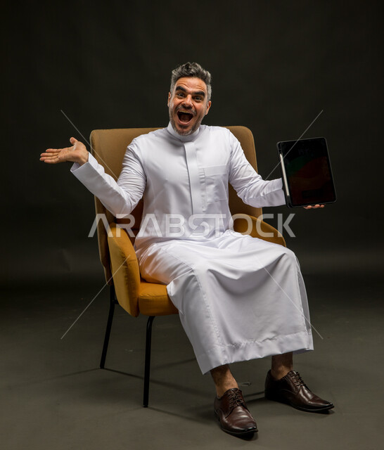 التسويق الرقمي الالكتروني، عرض شاشة إعلانية سوداء فارغة، استخدام الأجهزة والتقنيات الحديثة، رفع اليد للاعلى بإيماءات الانفعال والاندهاش بتطبيقات الجهاز اللوحي، بورتريه لشاب عربي خليجي سعودي يرتدي الزي التقليدي يمسك التابلت بتعابير الذهول، خلفية سوداء