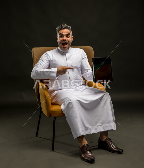 Using modern devices and technologies, electronic digital marketing, displaying a blank black advertising screen, pointing with the index finger with gestures of excitement and amazement at the tablet applications, a portrait of a Saudi Gulf Arab young man wearing traditional clothing holding the tablet with an astonished expression, a black background