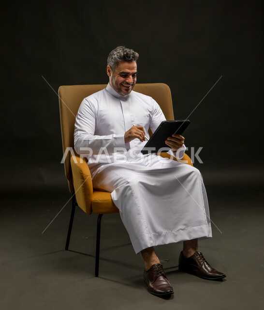 التواصل مع الأهل والاصدقاء عبر الانترنت، الضحك أثناء قراءة النكت والفكاهات، بورتريه لشاب عربي خليجي سعودي مبتسم يرتدي الزي التقليدي يقوم بمتابعة آخر أخبار وسائل التواصل الاجتماعي عبر الجوال، تصفح تطبيقات الهاتف المحمول، خلفية سوداء