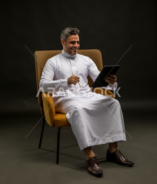 التواصل مع الأهل والاصدقاء عبر الانترنت، الضحك أثناء قراءة النكت والفكاهات، بورتريه لشاب عربي خليجي سعودي مبتسم يرتدي الزي التقليدي يقوم بمتابعة آخر أخبار وسائل التواصل الاجتماعي عبر الجوال، تصفح تطبيقات الهاتف المحمول، خلفية سوداء