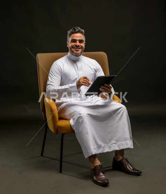 تصفح تطبيقات الهاتف المحمول، التواصل مع الأهل والاصدقاء عبر الانترنت، بورتريه لشاب عربي خليجي سعودي مبتسم يرتدي الزي التقليدي ينظر إلى الكاميرا بإيماءات السرور، متابعة آخر أخبار وسائل التواصل الاجتماعي عبر الجوال، خلفية سوداء