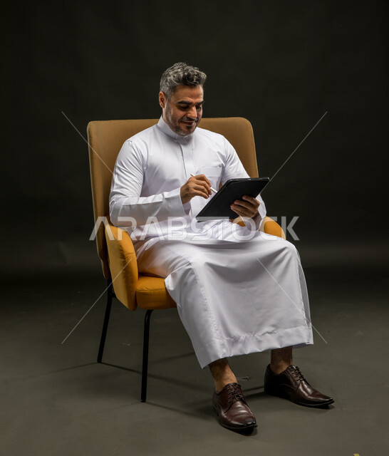 Browsing mobile applications and websites, communicating with family and friends via the Internet, a portrait of a smiling Saudi Gulf Arab young man wearing traditional clothing, following the latest social media news via mobile phone with happy gestures, black background.
