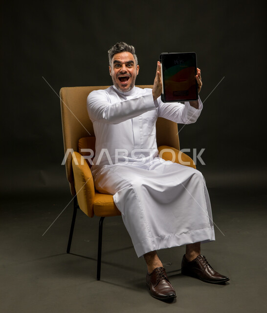 النظر إلى الكاميرا بإيماءات الدهشة والذهول والإعجاب، استخدام الأجهزة والتقنيات الحديثة، الإعلان والتسويق الرقمي الالكتروني للمنتجات الخليجية، عرض شاشة إعلانية سوداء فارغة، بورتريه لشاب عربي خليجي سعودي يرتدي الزي التقليدي يمسك  بيدهي جهاز التابلت، خلفية سوداء