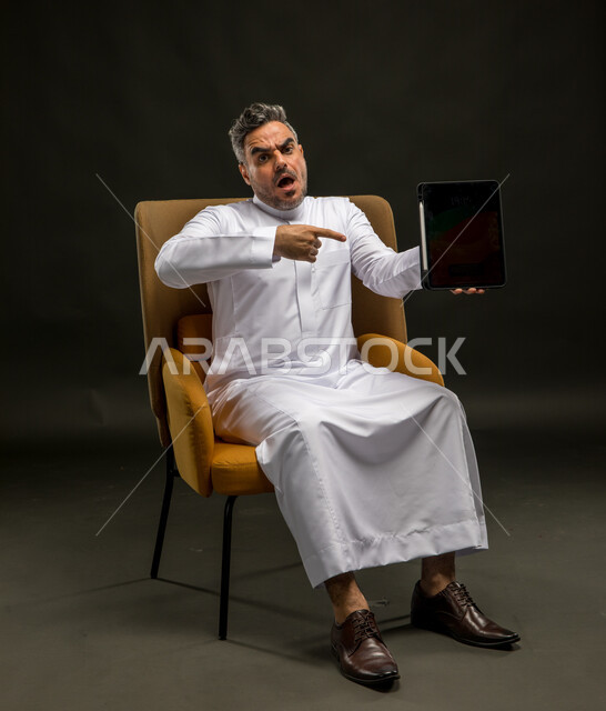 الإشارة إلى الجها اللوحي بتعابير الغضب والانزعاج، تصفح وسائل التواصل الاجتماعي، استخدام التقنيات الحديثة والمتطورة، بورتريه لشاب عربي خليجي سعودي يرتدي الثوب التقليدي يجلس على كرسي يحمل التابلت بتعابير عدم الرضا، خلفية سوداء
