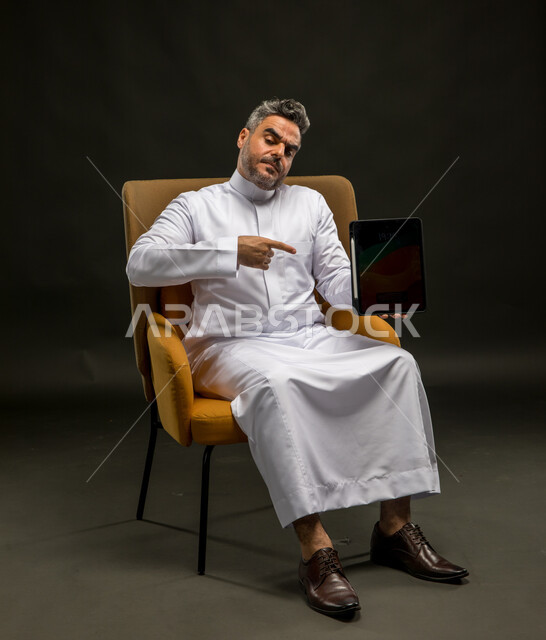 الإشارة إلى الجهاز اللوحي بتعابير الغضب والانزعاج، تصفح وسائل التواصل الاجتماعي، استخدام التقنيات الحديثة والمتطورة، بورتريه لشاب عربي خليجي سعودي يرتدي الثوب التقليدي يجلس على كرسي يحمل التابلت بتعابير عدم الرضا، خلفية سوداء