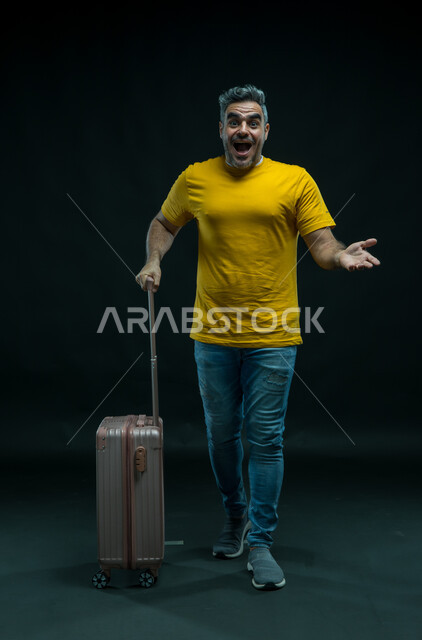 الإستعداد للرحلة السياحية الصيفيه، إيماءات الاندهاش والذهول, السياحة في إجازة الصيف, بورتريه لرجل عربي سعودي خليجي يرتدي الزي الكاجوال يمسك بيده حقيبة السفر, النظر إلى الكاميرا والشعور بالحماس والفرح، صورة الجسم بالطول الكامل, خلفية سوداء.