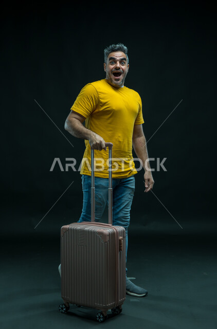 إيماءات الذهول والاندهاش, السياحة في إجازة الصيف, بورتريه لرجل عربي سعودي خليجي يرتدي الزي الكاجوال يمسك حقيبة السفر, الاستعداد لقضاء رحلة صيفية ممتعة, النظر إلى الكاميرا والشعور بالحماس والفرح، صورة الجسم بالطول الكامل, خلفية سوداء.