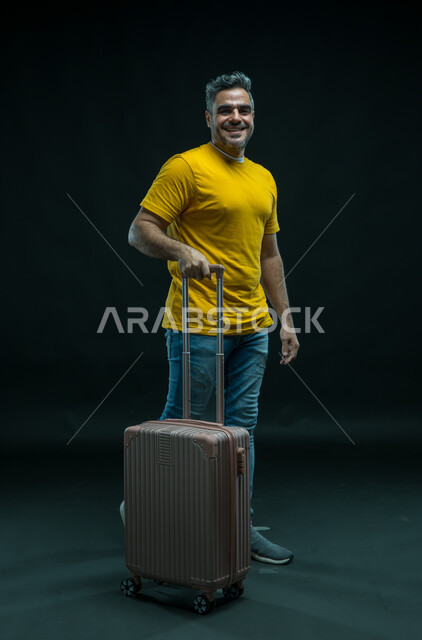 الاستعداد لقضاء رحلة صيفية ممتعة, السياحة في إجازة الصيف, إيماءات السرور والسعادة, بورتريه لرجل عربي سعودي خليجي مبتسم يرتدي الزي الكاجوال ينظر إلى الكاميرا و يمسك بيده حقيبة السفر, الشعور بالحماس والفرح، صورة للجسم بالكامل, خلفية سوداء.