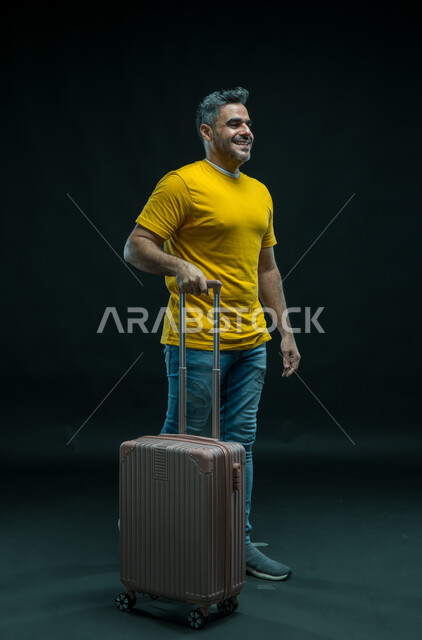 ايماءات المشي و الاستعداد للانطلاق وبدء الرحلة، الاستمتاع في قضاء إجازة الصيف، بورتريه لرجل عربي خليجي سعودي مبتسم يرتدي الزي الكاجوال يمسك بيده حقيبة السفر, الحماس للذهاب في جولة سياحية، صورة الجسم بالطول الكامل ، خلفية سوداء