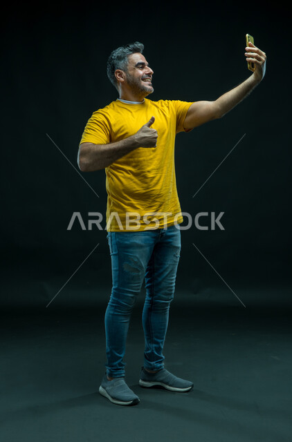 Quality sign gestures, taking souvenir selfies via mobile phone, using modern technologies, portrait of a Saudi Gulf Arab young man wearing casual clothes and documenting happy moments, making a video call via social media applications and websites, full-length body photo, black background