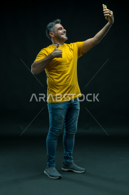  اجراء مكالمة فيديو عبر تطبيقات السوشال ميديا, ايماءات علامة الجودة، التقاط صور سيلفي تذكارية عبر الجوال, استخدام التقنيات الحديثة, بورتريه لشاب عربي خليجي سعودي يرتدي الزي الكاجوال ويقوم بتوثيق اللحظات السعيده, صورة الجسم بالطول الكامل, خلفية سوداء