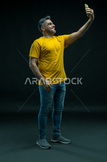 التواصل مع الاصدقاء والاقارب، اجراء مكالمة فيديو عبر تطبيقات السوشال ميديا, التقاط صور سيلفي تذكارية عبر الجوال, استخدام التقنيات الحديثة, بورتريه لشاب عربي خليجي سعودي يرتدي الزي الكاجوال ويقوم بتوثيق اللحظات السعيده, صورة الجسم بالطول الكامل, خلفية سوداء