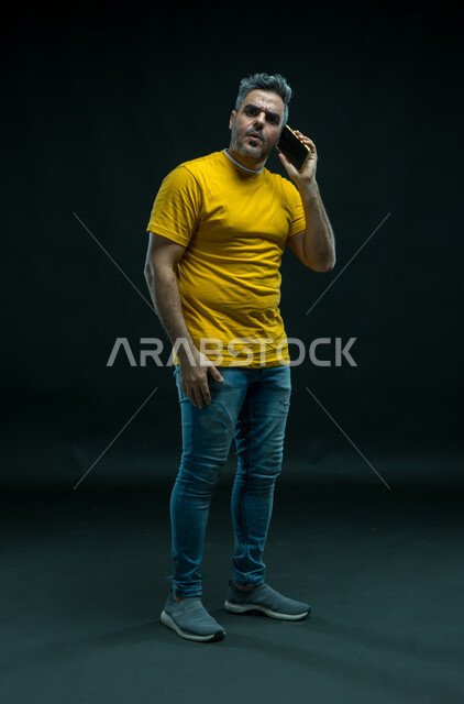 النظر إلى الكاميرا، الشعور بالانزعاج لسماع الأخبار السيئة, الاستفسار والسؤال عن أمر ما بإنفعالات وتعابير الغضب والقلق, بورتريه لشاب عربي خليجي سعودي يرتدي الزي الكاجوال يقوم بإجراء مكالمة هاتفية عبر الجوال ،صورة الجسم بالطول الكامل، خلفية سوداء