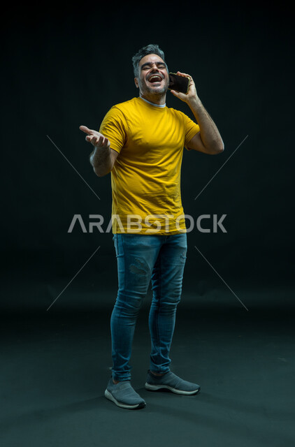 النظر إلى الكاميرا، إيماءات الفرح والسعاده, إجراء مكالمات صوتية عبر الهاتف المحمول, بورتريه لرجل عربي سعودي خليجي مسرور يرتدي الزي الكاجوال يقف باستقامة ويقوم بتبادل الاحاديث مع الاهل, الضحك والتسامر مع الأصدقاء، صورة الجسم بالكامل, خلفية سوداء