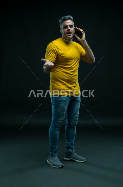 السؤال والاستفسار عن أمر ما بإنفعالات وتعابير الغضب والقلق, الشعور بالانزعاج لسماع الأخبار السيئة, بورتريه لشاب عربي خليجي سعودي يرتدي الزي الكاجوال يقوم بإجراء مكالمة هاتفية عبر الجوال وينظر إلى الكاميرا ،صورة الجسم بالطول الكامل، خلفية سوداء