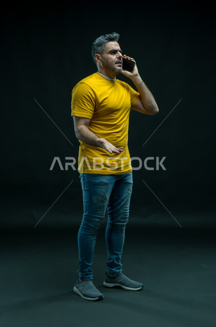Feeling upset to hear bad news, inquiring and asking about something with emotions and expressions of anger and anxiety, portrait of a Saudi Gulf Arab young man wearing a casual outfit making a mobile phone call, full length body photo, black background