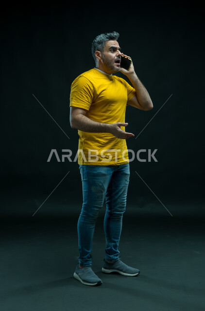 الشعور بالانزعاج لسماع الأخبار السيئة الصادمة ، بورتريه لشاب عربي خليجي سعودي يرتدي الزي الكاجوال يقوم بإجراء مكالمة هاتفية عبر الجوال بتعابير الاستغراب والاندهاش، استخدام التقنيات الحديثه المتطورة، صورة الجسم بالطول الكامل، خلفيه سوداء