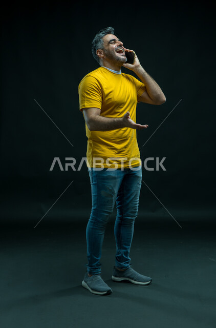 إيماءات الفرح والسعادة, إجراء مكالمات صوتية عبر الهاتف المحمول, بورتريه لرجل عربي سعودي خليجي مسرور يرتدي الزي الكاجوال يقف باستقامة ويقوم بتبادل الاحاديث مع الاهل, الضحك والتسامر مع الأصدقاء، صورة الجسم بالكامل, خلفية سوداء