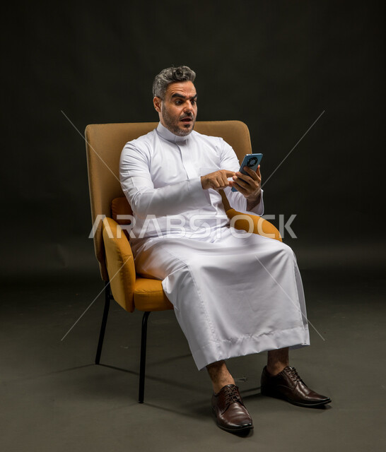 مشاهدة خبر غير سار على مواقع التواصل الاجتماعي، الخسارة في الصفقات المالية، استخدام أجهزة التواصل الحديثة والمتطورة, بورتريه لرجل عربي خليجي سعودي يرتدي الزي التقليدي يقوم بتصفح تطبيقات الهاتف المحمول بإيماءات الصدمة والعبوس, خلفية سوداء