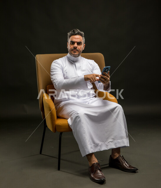 Looking at the camera with gestures of annoyance and frowning, watching unpleasant news on social networking sites, losing in financial deals, using modern and advanced communication devices, portrait of a Saudi Gulf Arab man wearing traditional clothing browsing mobile phone applications, black background