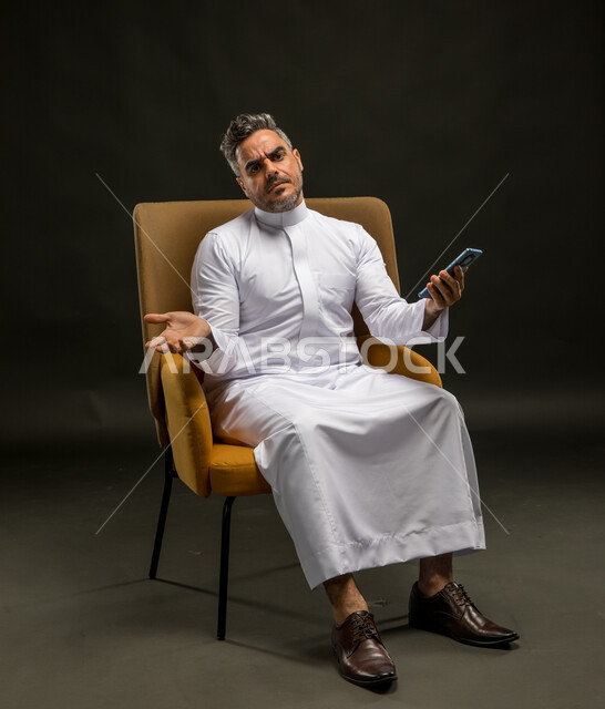 التفكير في حل لمشكلة ما، النظر إلى الكاميرا بإيماءات الحيرة والقلق والتوتر، مشاهدة خبر غير سار على مواقع التواصل الاجتماعي، استخدام الأجهزة الحديثة والمتطورة, بورتريه لرجل عربي خليجي سعودي يرتدي الزي التقليدي يقوم بتصفح تطبيقات الهاتف المحمول، خلفية سوداء