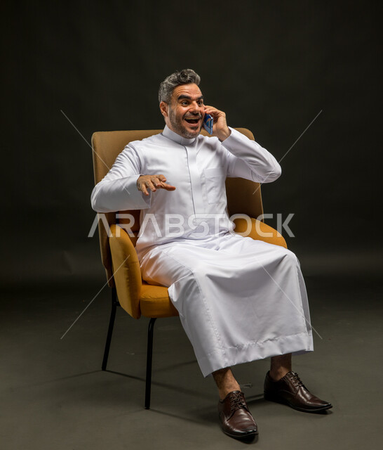 الضحك أثناء سماع النكات والفكاهات، تبادل الاحاديث المسلية والحوارات، التسامر مع الأصدقاء والأقارب بإيماءات السعادة، بورتريه لرجل عربي خليجي سعودي يرتدي الثوب التقليدي يقوم بإجراء مكالمة صوتيه عبر الهاتف المحمول، خلفية سوداء