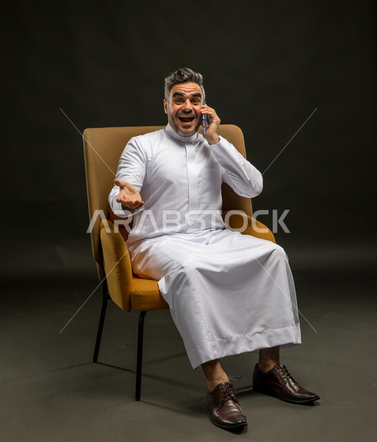 الضحك أثناء سماع النكات والفكاهات، تبادل الاحاديث المسلية والحوارات، التسامر مع الأصدقاء والأقارب بإيماءات السعادة، بورتريه لرجل عربي خليجي سعودي يرتدي الثوب التقليدي يقوم بإجراء مكالمة صوتيه عبر الهاتف المحمول، خلفية سوداء