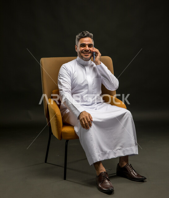 سماع الأخبار السارة والمفرحة، تبادل الاحاديث المسلية والحوارات، التسامر مع الأصدقاء والأقارب بإيماءات السعادة، بورتريه لرجل عربي خليجي سعودي يرتدي الثوب التقليدي يقوم بإجراء مكالمة صوتيه عبر الهاتف المحمول، خلفية سوداء