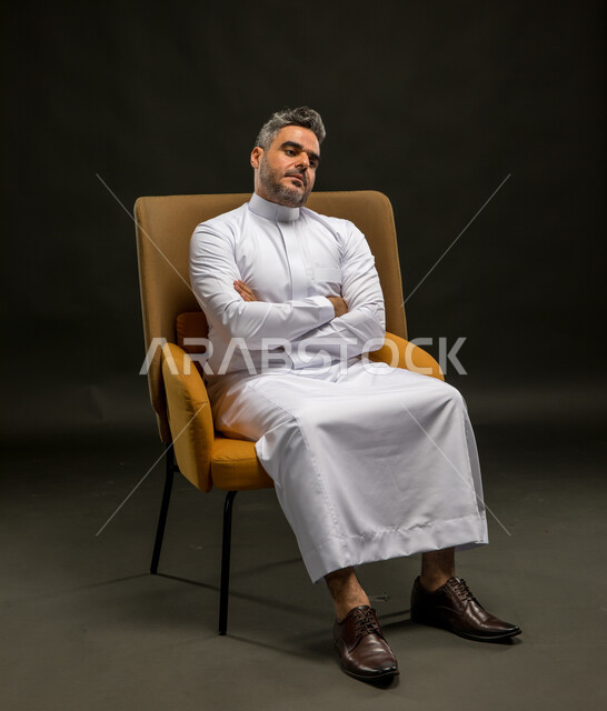 الشعور بالملل والتعب من ضغوطات العمل والحياة، تعابير التأمل وشرود الذهن، بورتريه لرجل عربي خليجي سعودي يرتدي الثوب التقليدي يجلس على الكرسي بأيدي متقاطعة وإيماءات التعب والارهاق، خلفية سوداء