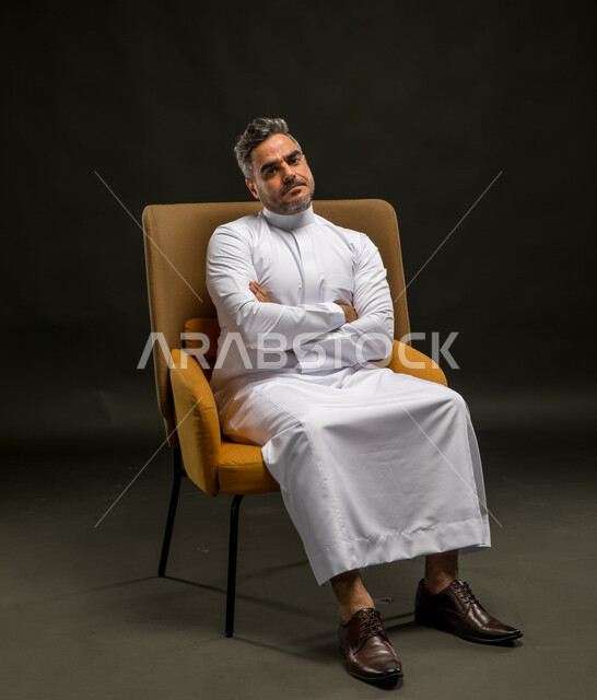 النظر إلى الكاميرا بتعابير الحزن والانكسار، الشعور بالملل والتعب من ضغوطات العمل والحياة، بورتريه لرجل عربي خليجي سعودي يرتدي الثوب التقليدي يجلس على الكرسي بأيدي متقاطعة وإيماءات التعب والارهاق، خلفية سوداء