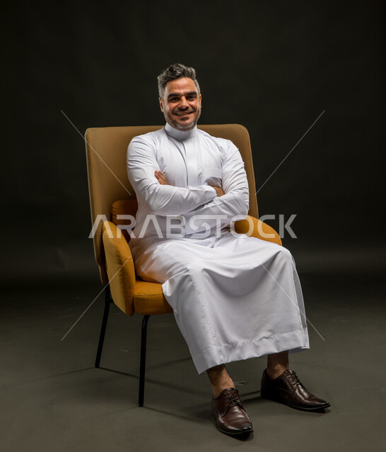 مفهوم الاهتمام بالمظهر الخارجي والرجولة والأناقة، بورتريه لرجل عربي خليجي سعودي مبتسم يرتدي الثوب التقليدي يجلس على الكرسي بأيدي متقاطعة ينظر إلى الكاميرا بإيماءات البشاشة والسرور، خلفية سوداء.
