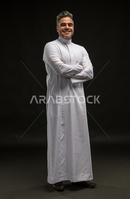 صورة شخصية للوثائق الرسمية، مفهوم الاهتمام بالمظهر الخارجي والرجولة والأناقة، بورتريه لرجل عربي خليجي سعودي مبتسم يرتدي الثوب التقليدي يقف بإستقامة وأيدي متقاطعة ينظر إلى الكاميرا بإيماءات البشاشة والسرور، صورة الجسم بالكامل، خلفية سوداء