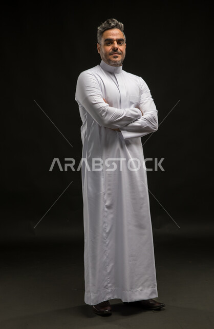 The concept of caring for external appearance, masculinity and elegance, a personal photo for official documents, a portrait of a smiling Saudi Gulf Arab man wearing a traditional dress, standing straight with crossed hands looking at the camera with gestures of joy and happiness, full body photo, black background