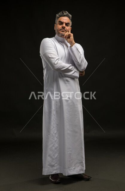 Thinking to solve a problem, facing problems and challenges in practical life, expressions of frowning and annoyance, portrait of a Saudi Gulf Arab man wearing a traditional dress, standing up straight, looking at the camera with gestures of disapproval and satisfaction, full body image, black background