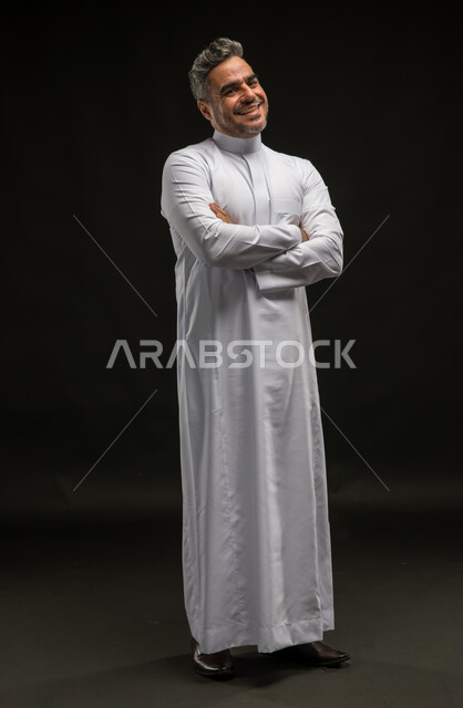 مفهوم الرجولة والأناقة، بورتريه لرجل عربي خليجي سعودي مبتسم يرتدي الثوب التقليدي يقف بإستقامة وأيدي متقاطعة ينظر إلى الكاميرا بإيماءات البشاشة والسرور، الاهتمام بالمظهر الخارجي، صورة الجسم بالطول الكامل، خلفية سوداء