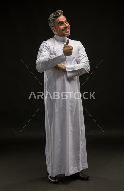 الشعور بالموافقه والقبول، إيماءات الدهشة والإعجاب والرضا, بورتريه لرجل عربي سعودي خليجي مبتسم يرتدي الثوب التقليدي يرفع إصبع الابهام نحو الأعلى بعلامة الجودة، مفهوم الإمتياز والتميز, صورة الجسم بالطول الكامل، خلفية سوداء