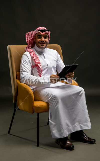 The concept of managing projects remotely, looking at the camera, gestures and expressions of joy and pleasure, using modern technical devices, a portrait of a Saudi Gulf Arab man wearing a shemagh and traditional thobe, sitting on a chair, completing work tasks via a tablet, black background